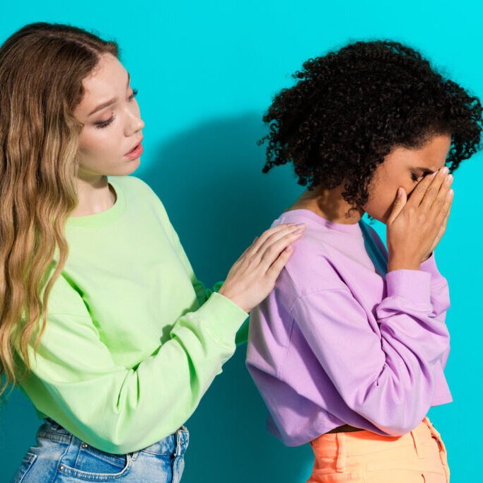 Stylish girlfriends in casual sportswear stand together by a vivid teal background, one offering a supportive gesture as the other expresses emotion.